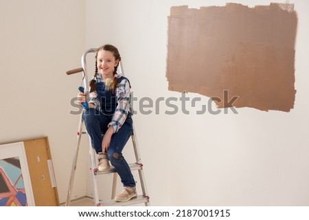 A very cute little girl in a denim suit and a checkered shirt sits on a ladder, holding a paintbrush with brown paint in her right hand. The child is very happy that he can help his parents.