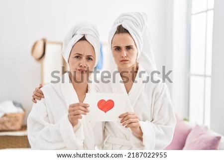 Middle age woman and daughter wearing bath robe holding heart card skeptic and nervous, frowning upset because of problem. negative person. 