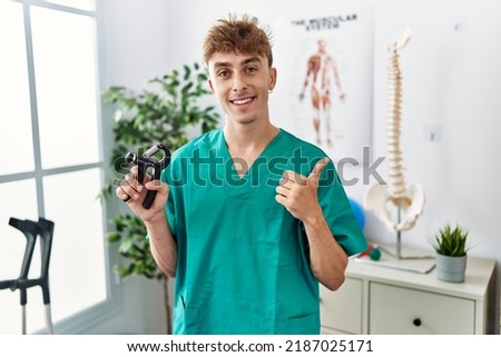Young caucasian physio man holding hand grip to train muscle at the clinic smiling happy and positive, thumb up doing excellent and approval sign 