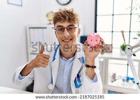 Young caucasian doctor man wearing doctor uniform holding piggy bank at the clinic smiling happy and positive, thumb up doing excellent and approval sign 