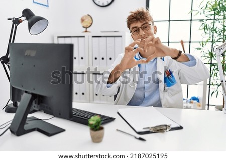 Young caucasian doctor man working at the clinic smiling in love doing heart symbol shape with hands. romantic concept. 