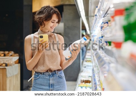 Young woman wear casual clothes backpack shopping at supermaket store buy choose dairy produce take milk read shelf life using mobile cell phone inside hypermarket Purchasing gastronomy food concept.