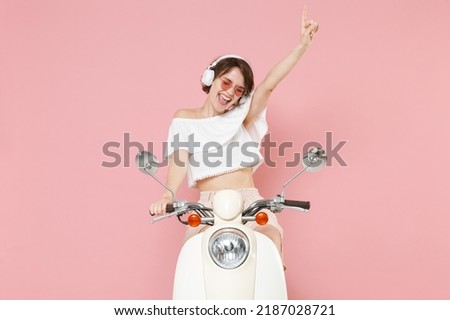 Cheerful young woman 20s in white summer clothes hat glasses listen music radio with headphones dance vibe point index finger up driving moped isolated on pastel pink colour background studio portrait