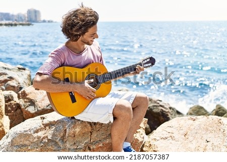 Young hispanic man playing classical guitar sitting on rock at the beach.