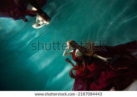 Photo underwater, a young beautiful woman in red with red hair reaches for the surface of the water, a human and his reflection. mystical underwater portrait