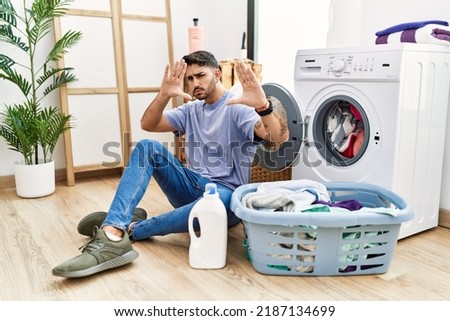 Young hispanic man putting dirty laundry into washing machine doing frame using hands palms and fingers, camera perspective 