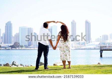 Adorable young couple holds hands and forms a heart towards the skyline of San Diego from across the bay                               