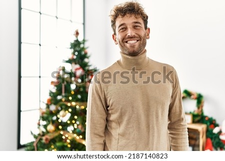 Young hispanic man smiling confident standing by christmas tree at home