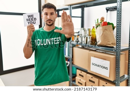Young hispanic man wearing volunteer t shirt holding we need you banner with open hand doing stop sign with serious and confident expression, defense gesture 
