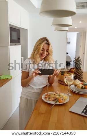 Blonde young woman taking photo of plate with shrimps, pasta and white wine. Food blogger. 