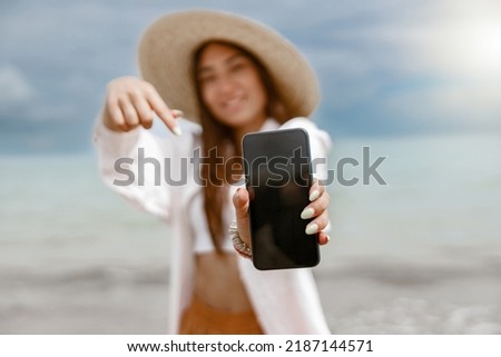 Smiling woman traveler in straw hat showing on phone standing on background of ocean