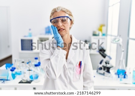 Middle age blonde woman working at scientist laboratory touching mouth with hand with painful expression because of toothache or dental illness on teeth. dentist 