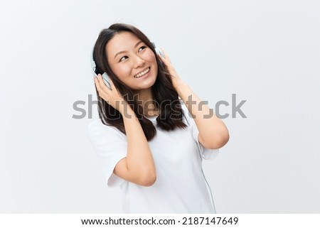 Dreaming happy cute Asian young female in white basic t-shirt touch headphones looks aside posing isolated on over white studio background. The best offer for ad. Favorite Music App Ad concept