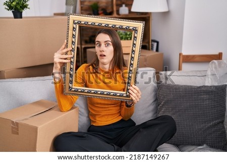 Young caucasian woman at new home holding empty frame in shock face, looking skeptical and sarcastic, surprised with open mouth 