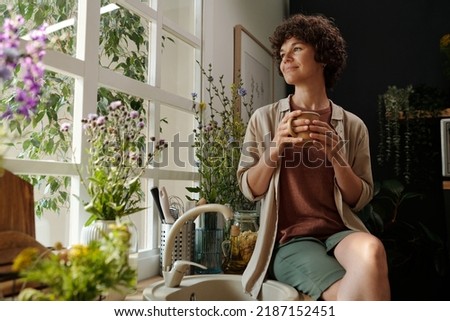 Young serene brunette with cup of coffee looking through window surrounded by plants while sitting by sink in the kitchen Royalty-Free Stock Photo #2187152451