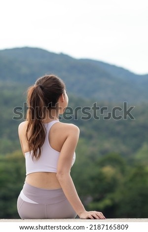 long haired woman in sports bra sits alone on roadside on dam crest against blurred background of green nature alone after jogging.  young woman sits alone on blurry background and has Copy Space Royalty-Free Stock Photo #2187165809