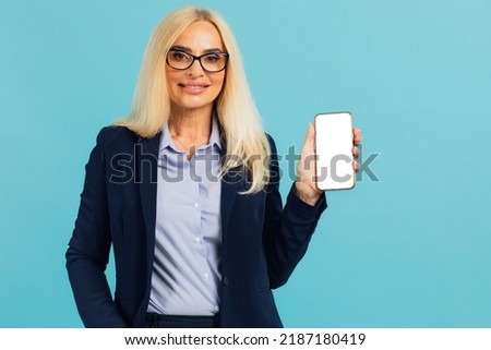 Portrait of smiling mature businesswoman in formal wear suit holding smartphone with white blank device screen in hand close up to camera, blue studio. Gadget with free space for mock up