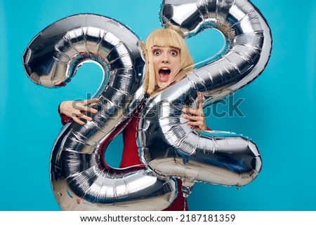 surprised, shocked woman in a red shirt stands with her mouth open on a blue background and holds inflatable balloons in the shape of the number twenty-two in silver color, hugging them with her