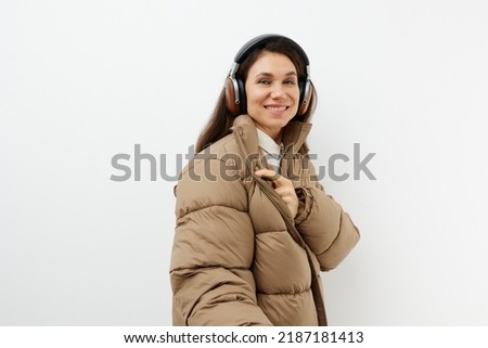 beautiful, happy, brunette listens to music in brown headphones, standing in a warm winter, stylish jacket and smiling cutely looking at the camera.