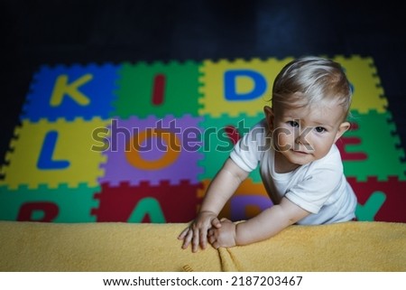 bird view of colorful kids puzzle mat playground in nursery with letters like Baby Love written on them lying on the floor while one year old blond baby in white body is playing