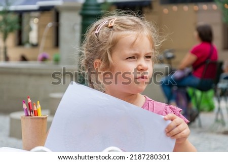 Street portrait of a little girl with a white sheet of paper in her hands on a blurry background of the old town.