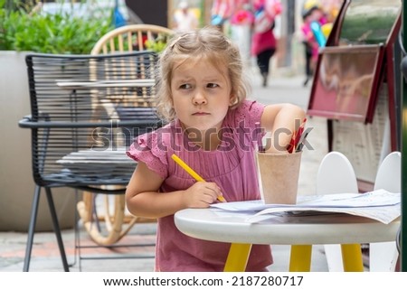 Street portrait of a beautiful 4-year-old girl drawing with colored pencils at a table in an outdoor cafe on a blurry background of the old town.