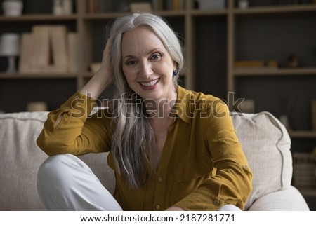 Happy joyful beautiful older 50s woman sitting on couch at home, looking at camera, laughing, smiling, touching long grey hair. Happy retirement, elderly age concept. Head shot portrait,