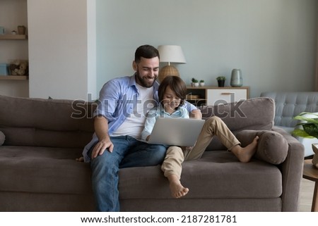 Dad and son sit on sofa enjoy weekend use laptop, rest at home spend leisure on internet, buying goods, make order on-line, choose cartoons or family movies on digital streaming services. Tech 