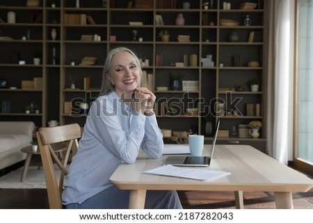 Happy cheerful elderly entrepreneur woman posing at home work table, sitting at laptop, looking at camera, smiling. Retired freelancer, blogger. Mature business woman portrait