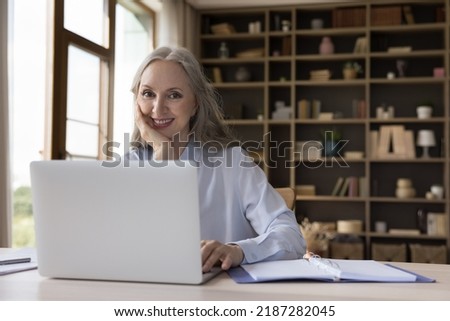 Happy mature freelance employee woman working at laptop, typing, sitting at desk with computer, notebook, looking at camera, smiling. Senior businesswoman at home workplace head shot portrait