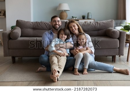 Couple with little children hugging sit on floor in living room smile look at camera feel happy, enjoy new house and weekend together at own first home. Bank loan, well-being family portrait concept Royalty-Free Stock Photo #2187282073