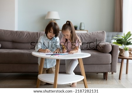 Beautiful preschoolers, little cute girl and boy sit at table at home draw pictures in sketchbook with coloured pencils, enjoy creative leisure spend pastime together look interested. Hobby concept