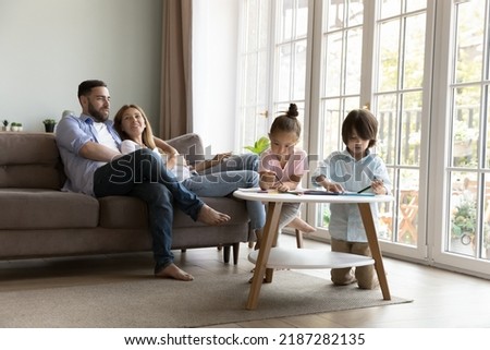 Young couple rest on comfort sofa their preschoolers children engaged in drawing holding colored pencils creating pictures in sketchbook, spend weekend together at home. Family pastime, hobby