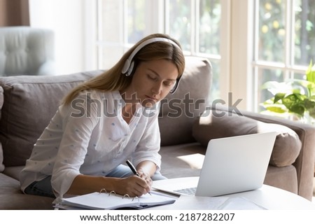 Focused busy woman jotting taking notes by work or study while listen information through headphones seated at table with laptop. E-learning, self-education. On-line class, modern technology concept