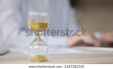 Sand watch object close up with employee human hands typing on laptop in blurred background. Hourglass with running sand on work table. Business, time management, deadline concept