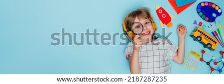 Funny girl with magnifying glass surrounded by different school stationery lying on blue background. Top view, flat lay. Back to school concept. Panorama. Horizontal banner with copy space. Royalty-Free Stock Photo #2187287235