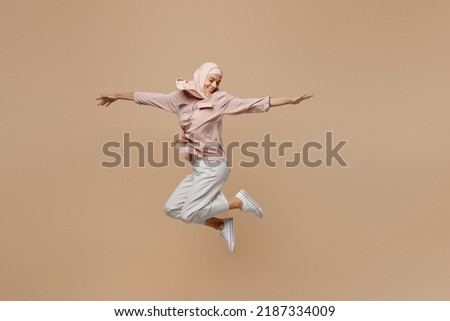 Full body young fun arabian asian muslim woman she wear abaya hijab pink clothes jump high with outstretched hands isolated on plain pastel light beige background. People uae islam religious concept Royalty-Free Stock Photo #2187334009