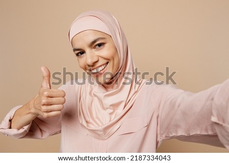 Close up young arabian asian muslim woman she in abaya hijab pink clothes do selfie shot pov mobile cell phone show thumb up isolated on plain beige background. People uae middle eastern islam concept