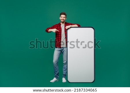 Full body young happy man he 20s in red shirt grey t-shirt point finger on big huge blank screen mobile cell phone with workspace copy space mockup area isolated on plain dark green background studio