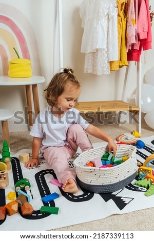 A little girl sitting on floor plays with toy wooden blocks on a rug with a picture of a road in playroom. Educational game for baby and toddler in modern nursery. 
