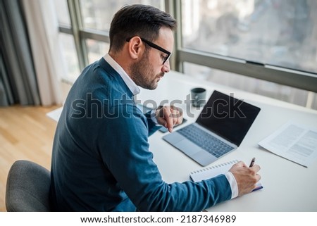 High angle view of serious professional writing notes in diary. Young businessman is using laptop while working at desk. He is wearing sweater in corporate office. Royalty-Free Stock Photo #2187346989