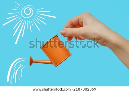 A woman's hand with a miniature orange watering can on a blue background. Close-up. Royalty-Free Stock Photo #2187382369