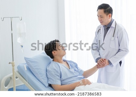 Senior doctor and young male patient who lie on the bed while checking pulse, consult and explain with nurse taking note and supporting in hospital wards. Royalty-Free Stock Photo #2187386583