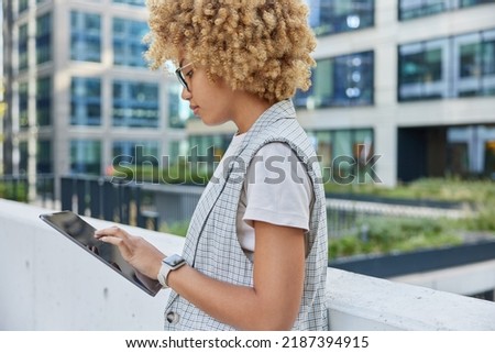 Half length shot of beautiful curly haired woman in elegant formal clothes satisfied with successful career reads text on her digital tablet during recreation time poses against office building Royalty-Free Stock Photo #2187394915