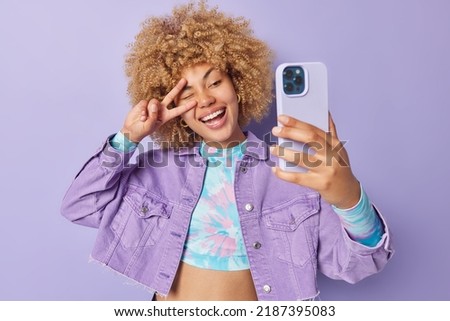 Glad positive European woman with curly blonde hair makes peace gesture over eye smiles happily takes selfie via smartphone wears stylish clothes poses against purple background enjoys life. Royalty-Free Stock Photo #2187395083