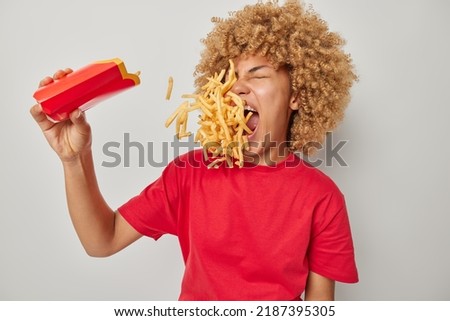 Cheat meal concept. Curly haired woman eats french fries containing much calories dressed in casual red t shirt being fast food lover has mouth full of chips poses indoor against grey background Royalty-Free Stock Photo #2187395305