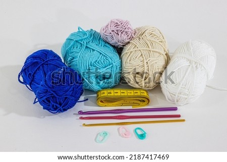 Cotton balls in blue, white, beige and pink colors next to a yellow measuring tape, pink, purple and gold metallic crochet hooks, pink and green stitch markers on a surface on a white background