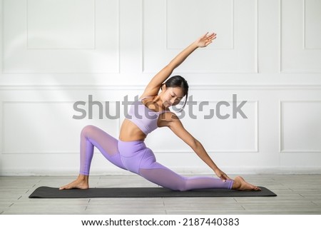 Side view of Asian woman wearing purple sportwear doing Yoga exercise in front of windows,Yoga low lunge pose or Anjaneyasana. Calm of healthy young woman breathing and meditation yoga at home Royalty-Free Stock Photo #2187440383
