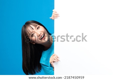 Portrait of smiling asian woman holding empty blank board on blue studio background. Happy young girl standing with white square paper card for text and showing card to camera.