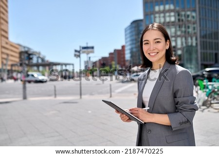 Young smiling Asian professional woman wearing suit holding digital tablet corporate digital information global technology standing on big city street using pad computer outside. Portrait Royalty-Free Stock Photo #2187470225